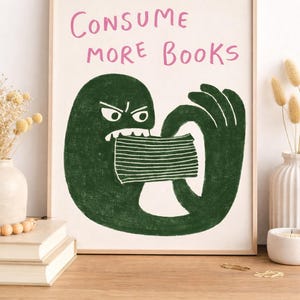 May include: A framed art print with the text "CONSUME MORE BOOKS" in pink. The illustration depicts a green monster-like figure biting a stack of books. The artwork has a whimsical and playful style, suitable for a home library or reading corner.