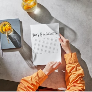 May include: A white paper with the text "Jen's Bachelorette" in cursive, with a word search puzzle below. The person is wearing an orange long-sleeve shirt. A glass of water with a gold straw and orange slice is on the left, and a glass of water with an orange slice is on the top.