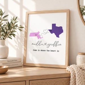 Puede incluir: Una lámina enmarcada con un mapa de Massachusetts en violeta claro y un mapa de Texas en violeta oscuro conectados por una trayectoria de vuelo punteada con un corazón. El texto dice "maddie & griffin" y "Home is where the heart is".