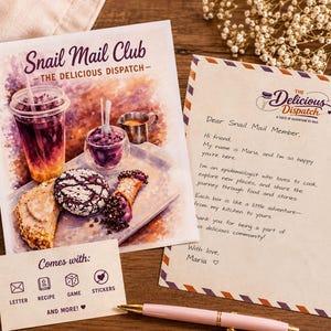 May include: A promotional image for "Snail Mail Club - The Delicious Dispatch" featuring a watercolor-style illustration of food and drinks. Includes a letter, recipe, game, and stickers. A pink pen rests on the wooden surface.