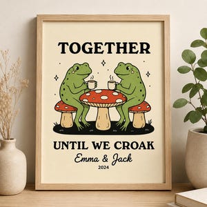 May include: A framed wall art print featuring two green frogs sitting on mushroom stools at a mushroom table, each holding a white mug. The text reads TOGETHER UNTIL WE CROAK, Emma & Jack, and 2024. The illustration is set against a cream background.