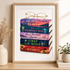 May include: A framed print featuring a stack of five books topped with gold-rimmed glasses. The book titles are The Night We Met, My Husband's Wife, Our Perfect Storm, First Lie Wins, and The Women, each with colorful, illustrative cover art.