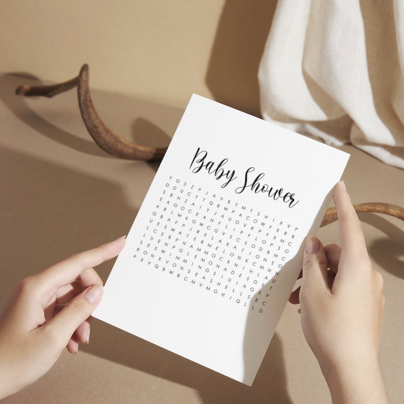 May include: A white paper with the words "Baby Shower" in elegant script. Below the title is a word search puzzle with words like "watermelon" and "seashells". The paper is held by two hands.