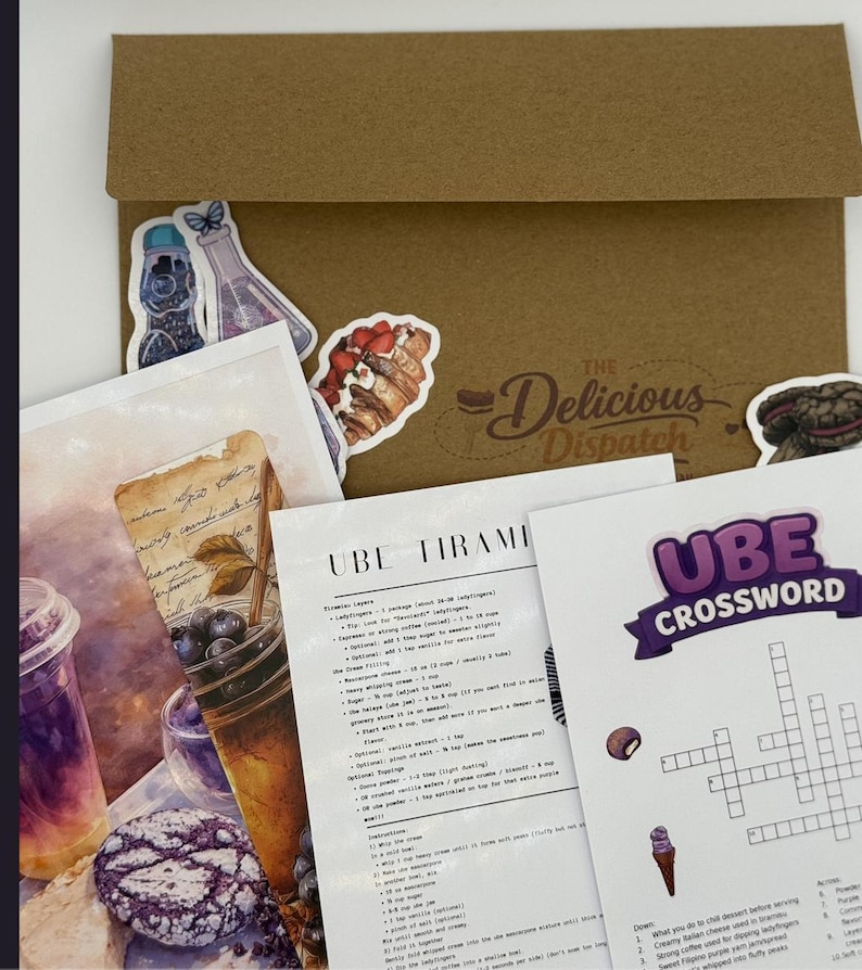 May include: A kraft paper envelope with stickers and printed recipe cards. Stickers include a butterfly, a beaker, and a pastry. Recipe cards feature text for Ube Tiramisu and an Ube crossword puzzle. The Delicious Dispatch logo is visible.