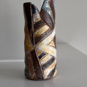 May include: A handmade ceramic vase with a unique, geometric design. The vase features a mix of colors, including brown, white, yellow, and blue, arranged in a textured pattern. The top has a distinctive, open shape. This decorative piece is suitable for home decor.