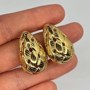 May include: A pair of gold-colored, woven-style earrings. The earrings have a teardrop shape with an open, basket-weave design. The earrings are displayed against a neutral background.