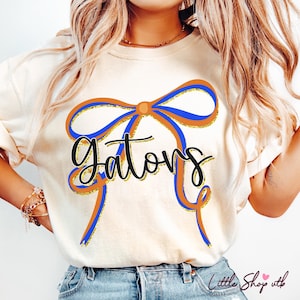 May include: Cream-colored t-shirt featuring a large bow design in orange, blue, and gold. The word "Gators" is written in a black script font across the bow. The shirt is a casual style.