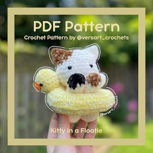 Puede incluir: Un gato de ganchillo en un flotador amarillo con un pato amarillo. El gato es blanco con manchas marrones y una nariz negra. El texto "PDF Pattern" y "Kitty in a Floatie" son visibles.
