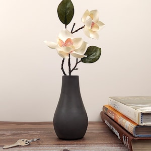 May include: A black ceramic vase with a pearlescent white magnolia flower arrangement. The vase is sitting on a wooden table with a stack of books.