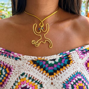 May include: A gold-colored wire necklace featuring a stylized human figure pendant with two small blue beads. The necklace is worn over a colorful, patterned top with a crochet design.