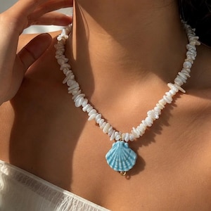 May include: A necklace featuring a strand of irregular white shell chips and a single light blue ceramic seashell pendant. The pendant is attached to the strand with a small gold-toned metal jump ring, creating a layered look around the neck.