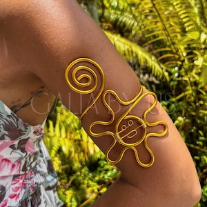 May include: A gold-colored arm cuff featuring a spiral design and a stylized sun shape. The cuff is made of wire and is worn on the upper arm. The background is blurred, with green foliage.