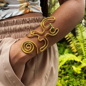 May include: A gold-colored arm cuff with a spiral design and abstract shapes. The cuff features small green beads and is worn on the arm. The background is blurred, with green foliage visible.