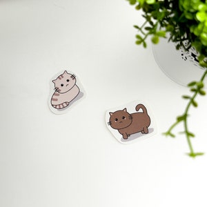 May include: Two cute cartoon cat stickers. One is a brown cat with a sad expression and the other is a light brown cat with a sad expression.
