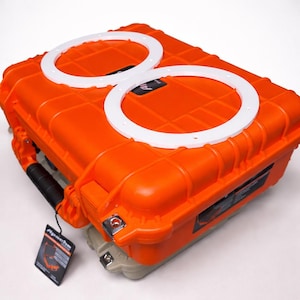 May include: An orange Apache hard case with two white circular inserts. The case has a textured surface, a black handle, and a tag. The case is designed to protect items during transport.