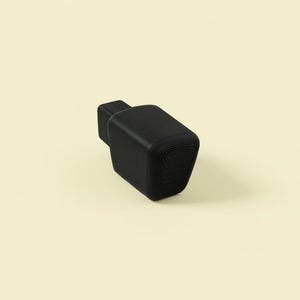 May include: A black, geometric bottle stopper with a rectangular body and a smaller, square top. The stopper has a textured, layered appearance and is set against a light beige background.