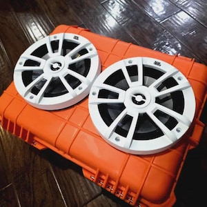 May include: Two white and black marine speakers with a radial design are placed on top of an orange, rectangular case. The speakers have a circular shape and are made of plastic. The case has a textured surface.