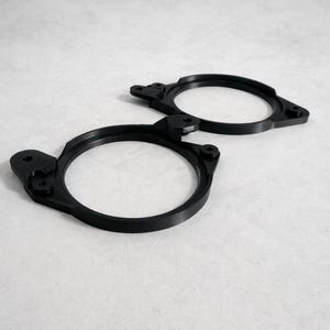 May include: Two black circular components with mounting features are shown against a white background. Each component has a central opening and several protruding sections with holes. The parts appear to be made of a rigid material.