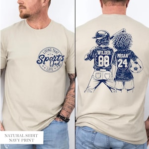 May include: A natural-coloured t-shirt with a navy blue graphic on the front that says "Living That Sports Dad Life." The back of the shirt features illustrations of a baseball player and a soccer player with names and numbers.