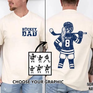 May include: Ivory-colored t-shirt with navy ink graphics. The front features "HOCKEY DAD" text. The back displays a hockey player with the name "RILEY" and the number "8". A graphic selection is shown below. The shirt is a unisex design.