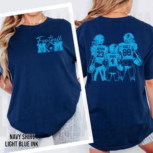 May include: A navy blue t-shirt featuring light blue graphics. The front displays the text "Football MOM" with a football icon. The back shows three football players in uniform with the names "KYEN 23", "KRUZ 4", and "KOLT 88" printed in light blue.