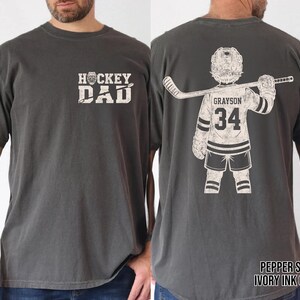 May include: Pepper-colored t-shirt with "HOCKEY DAD" text on the front. The back features a hockey player graphic with the name "Grayson" and the number "34". The shirt is made of a soft material.