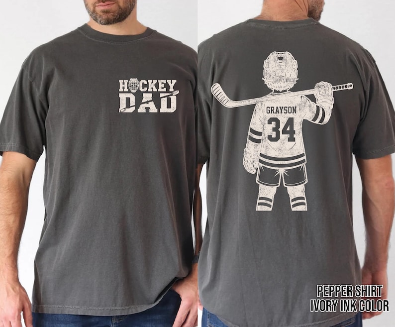 May include: Pepper-colored t-shirt with "HOCKEY DAD" text on the front. The back features a hockey player graphic with the name "Grayson" and the number "34". The shirt is made of a soft material.