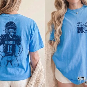 May include: A light blue t-shirt featuring a dark blue graphic on the back of a football player wearing a helmet and jersey with the name KLORMAN and number 11. The front chest area displays the text Football MOM with a football graphic.