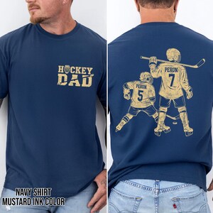 May include: Navy blue t-shirt with "HOCKEY DAD" in mustard yellow on the front. The back features a hockey player graphic with the name "PERUN" and the number "7" and "5". The shirt's color is navy with mustard yellow ink.
