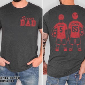 May include: Dark grey heather shirt with red ink text and graphics. The front of the shirt reads "Soccer Dad" in a bold, stylized font. The back features two soccer players with the names "Michael" and "Brayden" and their jersey numbers.
