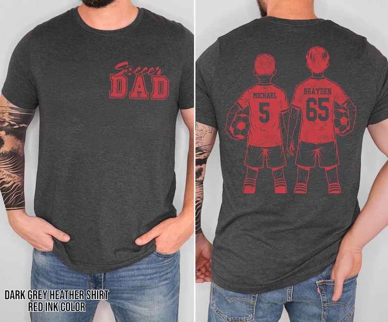 May include: Dark grey heather shirt with red ink text and graphics. The front of the shirt reads "Soccer Dad" in a bold, stylized font. The back features two soccer players with the names "Michael" and "Brayden" and their jersey numbers.