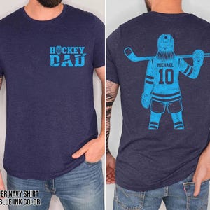 Pode incluir: T-shirt azul marinho mesclado com tinta azul clara. A frente da camisa tem o texto "HOCKEY DAD". A parte de tr&aacute;s da camisa apresenta um gr&aacute;fico de jogador de h&oacute;quei com o nome "MICHAEL" e o n&uacute;mero "10".