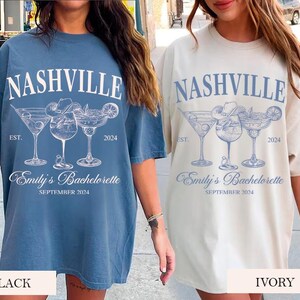 May include: Two oversized t-shirts with a blue and white design featuring the text "Nashville" and "Emily's Bachelorette" with a graphic of three cocktail glasses. The t-shirts are shown in black and ivory.