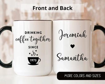 May include: Two white ceramic mugs with black handles and black text. The mug on the left says "Drinking coffee together since 1973" with a small coffee cup icon. The mug on the right says "Jeremiah Samantha" with a heart icon.