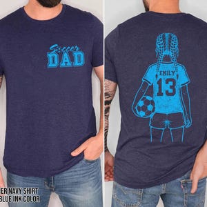 May include: Heather navy blue t-shirt with light blue ink. The front features the text "Soccer DAD". The back displays a light blue illustration of a girl holding a soccer ball, with the name "EMILY" and the number "13".