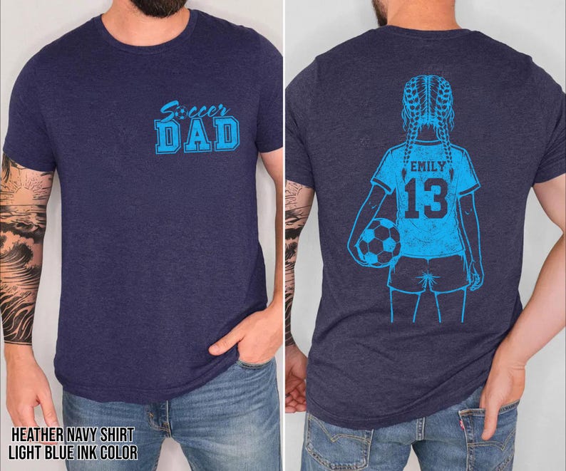 May include: Heather navy blue t-shirt with light blue ink. The front features the text "Soccer DAD". The back displays a light blue illustration of a girl holding a soccer ball, with the name "EMILY" and the number "13".
