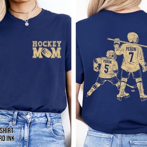 May include: Navy blue t-shirt with "HOCKEY MOM" in mustard yellow on the front. The back features a mustard yellow graphic of two hockey players with the text "PERUN" and numbers 5 and 7. The shirt is made of a soft material.