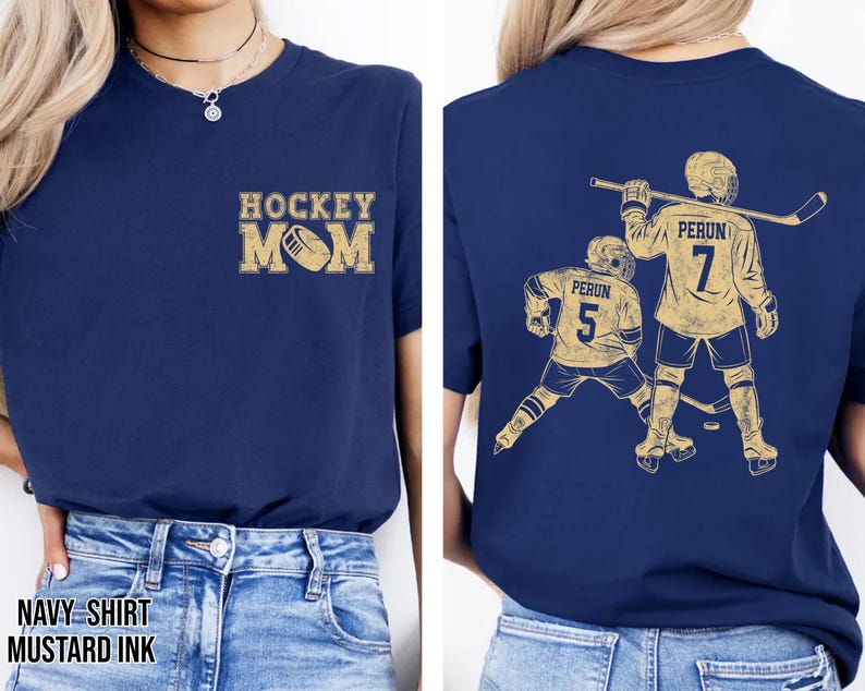 May include: Navy blue t-shirt with "HOCKEY MOM" in mustard yellow on the front. The back features a mustard yellow graphic of two hockey players with the text "PERUN" and numbers 5 and 7. The shirt is made of a soft material.