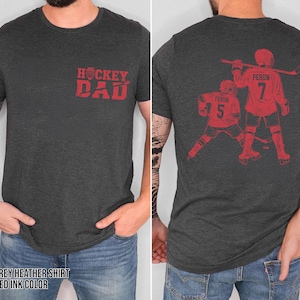 Pode incluir: T-shirt cinza escuro mesclado com tinta vermelha. A frente apresenta o texto "Hockey Dad". A parte de tr&aacute;s exibe um gr&aacute;fico de jogador de h&oacute;quei com o nome "Perun" e o n&uacute;mero "7".