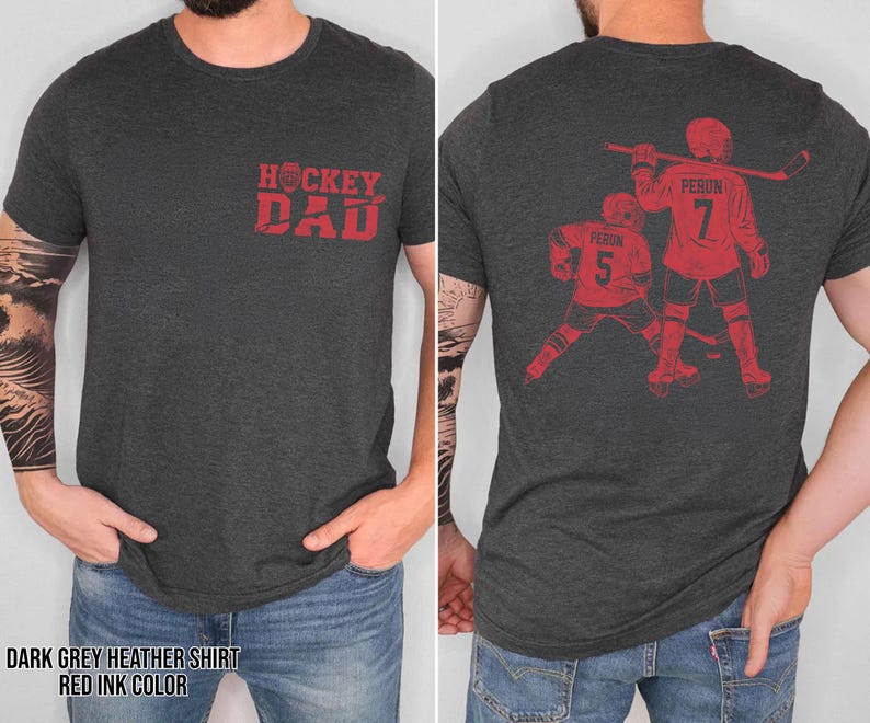 Pode incluir: T-shirt cinza escuro mesclado com tinta vermelha. A frente apresenta o texto "Hockey Dad". A parte de tr&aacute;s exibe um gr&aacute;fico de jogador de h&oacute;quei com o nome "Perun" e o n&uacute;mero "7".