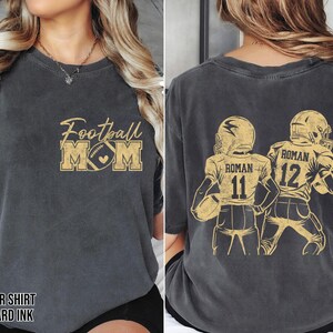 May include: A charcoal gray short-sleeve t-shirt with mustard yellow graphics. The front features the text Football MOM with a football icon. The back displays two football players in jerseys with the name ROMAN and numbers 11 and 12.