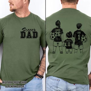 May include: Military green t-shirt with the text "Soccer Dad" in black on the front. The back of the shirt features a family of three holding soccer balls, with names and numbers. The shirt is made of a soft material.