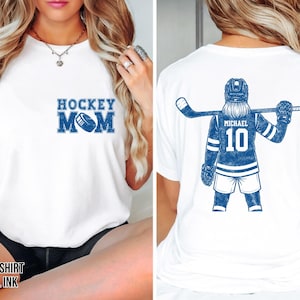 May include: White t-shirt with "HOCKEY MOM" in blue block letters and a hockey puck graphic. The back of the shirt features a blue hockey player graphic with the name "MICHAEL" and the number "10".
