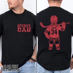 May include: Black t-shirt with red ink design. The front features the text "HOCKEY DAD". The back displays a hockey player graphic with the name "KATIE" and the number "34". The shirt is a classic crew neck style.