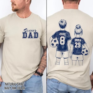 May include: A beige t-shirt with the text "Soccer Dad" in navy blue. The back of the shirt features illustrations of two soccer players, with the names "Zeke" and "Emma" and their jersey numbers. The shirt is labeled "Natural Shirt Navy Ink Color".
