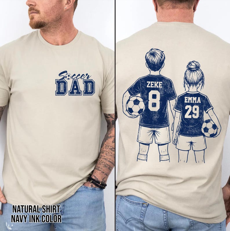 May include: A beige t-shirt with the text "Soccer Dad" in navy blue. The back of the shirt features illustrations of two soccer players, with the names "Zeke" and "Emma" and their jersey numbers. The shirt is labeled "Natural Shirt Navy Ink Color".