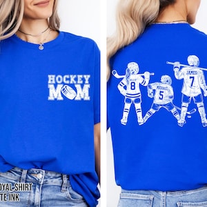 May include: Royal blue t-shirt with white text and graphics. The front features "HOCKEY MOM" with a hockey puck. The back displays three hockey players with names and numbers. The shirt's colour is described as "TRUE ROYAL SHIRT" with "WHITE INK".