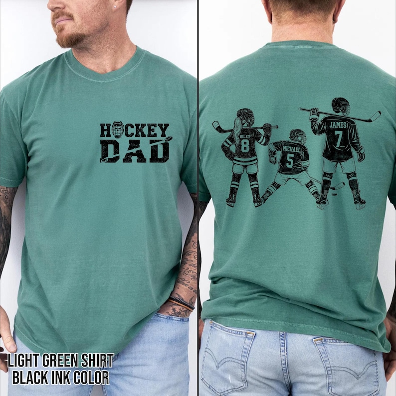 May include: Light green t-shirt with the text "HOCKEY DAD" in black on the front. The back of the shirt features a black graphic of three hockey players with names and numbers. The shirt is a comfortable, casual style.