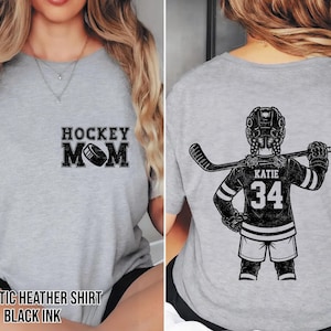 May include: Heather grey athletic shirt with "HOCKEY MOM" and a hockey puck graphic on the front. The back features a hockey player illustration with the name "KATIE" and the number "34". The shirt is printed with black ink.