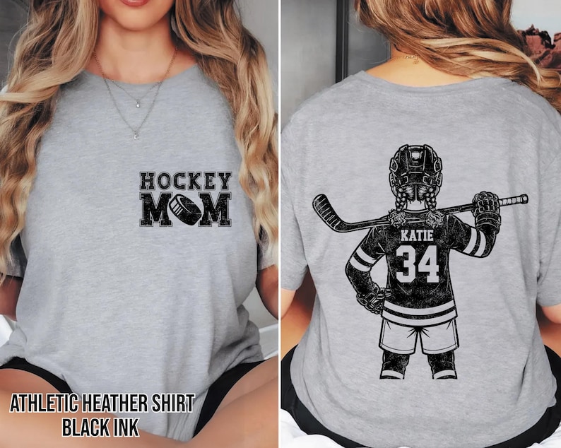 May include: Heather grey athletic shirt with "HOCKEY MOM" and a hockey puck graphic on the front. The back features a hockey player illustration with the name "KATIE" and the number "34". The shirt is printed with black ink.